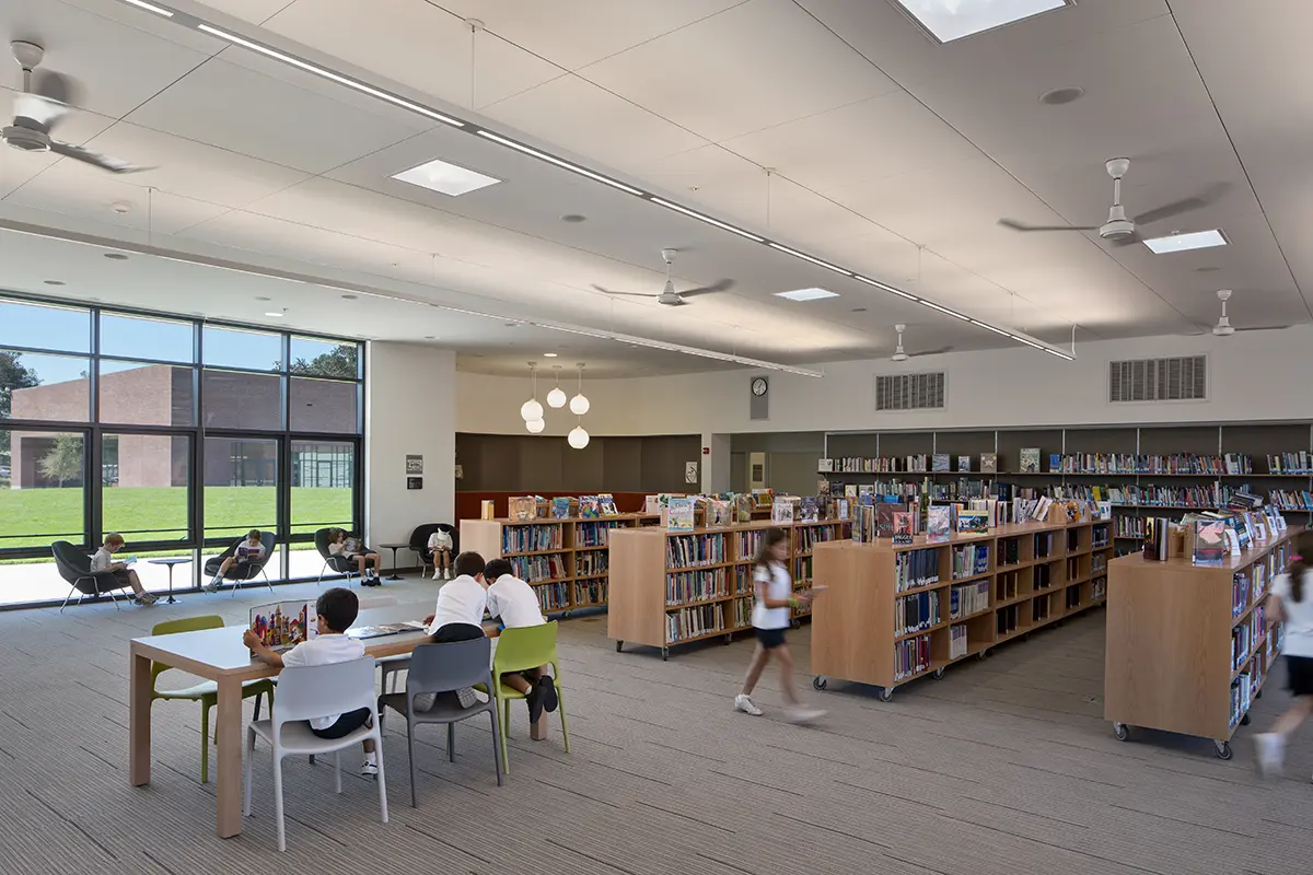 Modern primary school library with students reading at tables and shelves filled with books, large floor-to-ceiling windows, overhead lighting, and ceiling fans.